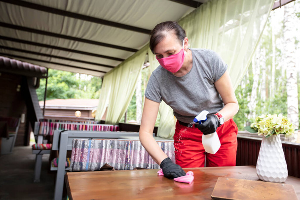 Cameriere indossando maschera protettiva durante la disinfezione tavoli al caffè all'aperto - Foto, immagini