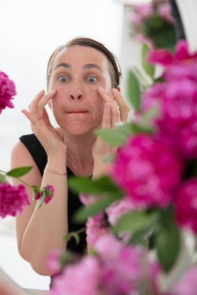 Eine Frau mittleren Alters untersucht Falten vor einem Hintergrund aus Blumen. Pflege für alternde Haut. - Foto, Bild