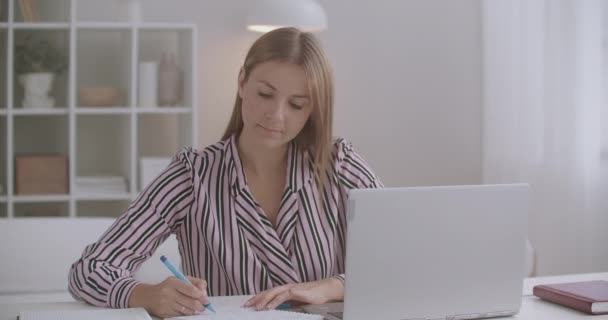 önképzés és e-learning, nő néz online képzés kijelzőjén laptop és írás papíron - Felvétel, videó