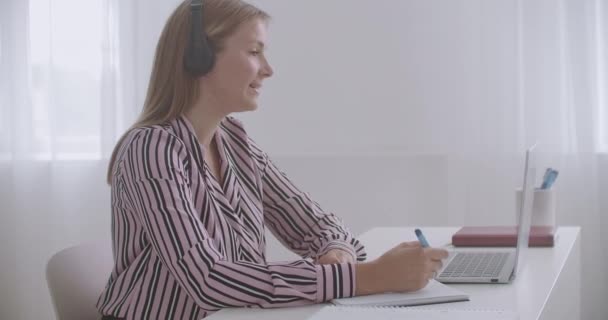 fiatal nő szakember kommunikál online video chat a laptopon, a fejhallgató, írás a copybook - Felvétel, videó