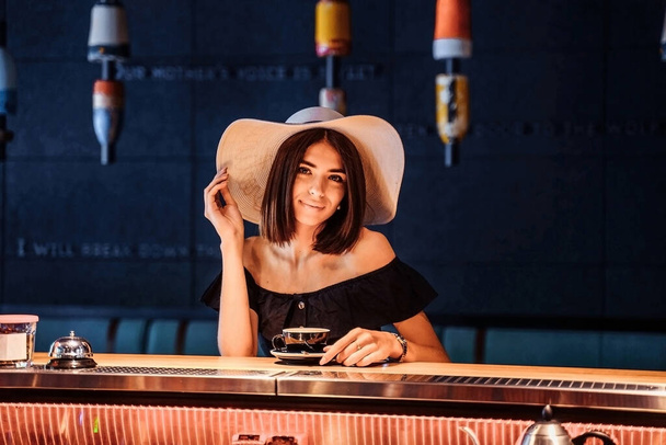 Portrait of a young woman drinking coffee at the bar in a cafe with a beautiful modern brown interior. A pretty girl who loves coffee is sitting in a restaurant. Holding a white hat with a hand. - Φωτογραφία, εικόνα