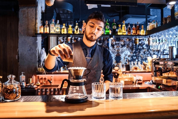 En una cafetería popular con luz suave y amarilla por la noche, un barista profesional vierte agua caliente de una tetera de metal en granos de café molidos. Una forma alternativa de hacer café. Filtro de café. - Foto, Imagen