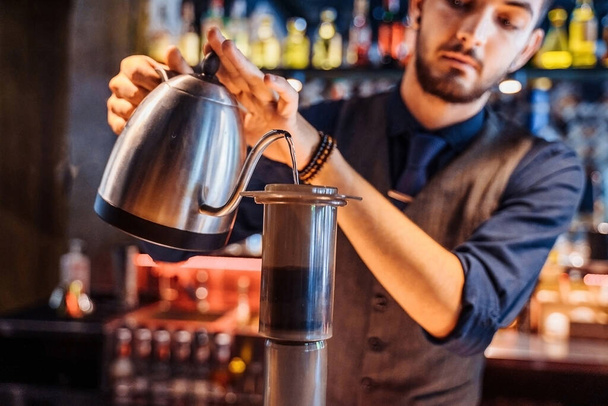 In een populair café met zacht, geel avondlicht giet een professionele barista heet water uit een metalen ketel in gemalen koffiebonen. Een alternatieve manier om koffie te zetten. Koffiefilter. - Foto, afbeelding