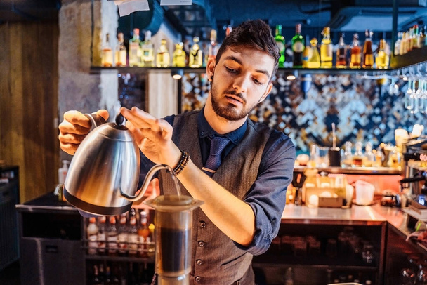 Em um café popular com luz suave e amarela, um barista profissional derramando água quente de uma chaleira de metal em grãos de café moídos. Uma forma alternativa de fazer café. Filtro de café. - Foto, Imagem