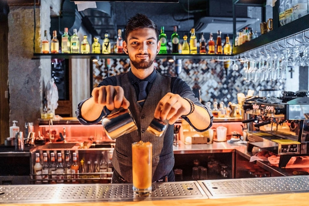Jóképű barna barista kelet-kaukázusi megjelenésű, italt készít a bárban egy étteremben. Keverés egy kávé és narancslé hozzáadásával jég a koktél. Szívószálat helyez be.. - Fotó, kép