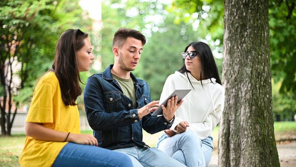 Three students studying together with a digital tablet sitting on a bench outdoor - 写真・画像