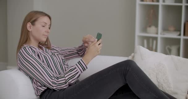 adult woman is browsing social nets in smartphone, lying on couch at home, swiping and scrolling - Materiaali, video