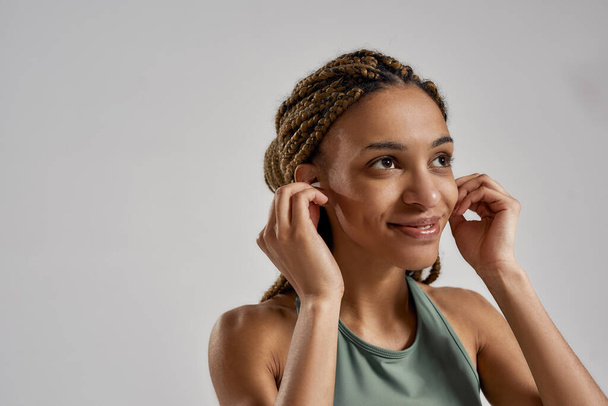 Music for workout. Portrait of young happy mixed race fitness woman wearing wireless earbuds and smiling while standing isolated over grey background - Foto, afbeelding