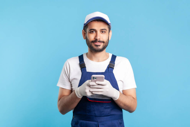 Handyman in overalls and gloves holding mobile phone, using cellphone messenger to accept online order as delivery, repair and maintenance services. indoor studio shot isolated on blue background - Photo, image