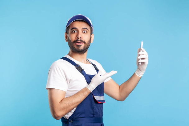 Surprised workman in overalls and gloves pointing mobile phone and looking amazed, shocked by online delivery order app, house repair maintenance services. studio shot isolated on blue background - Fotografie, Obrázek