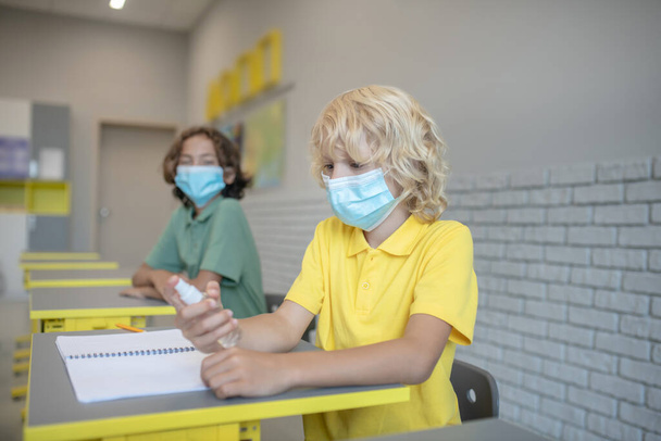 Een blonde jongen met een masker die ontsmettingsmiddel op z 'n handen spuit. - Foto, afbeelding