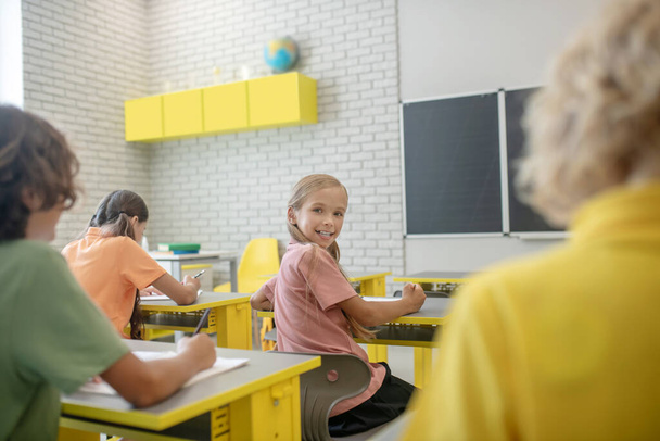 Schattig meisje zit aan het bureau in de klas en glimlacht naar haar klasgenoot - Foto, afbeelding