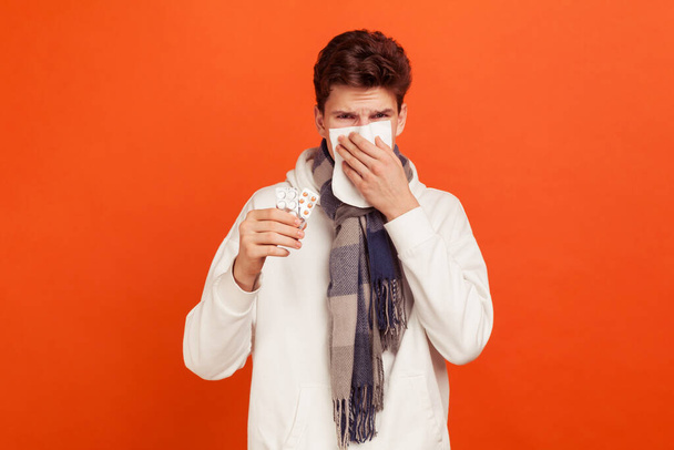 Sad unhealthy young man in casual sweater with hood and checkered scarf on neck holding tablet plates in hand, suffering from flu, runny nose. Indoor studio shot isolated on orange background - Photo, Image