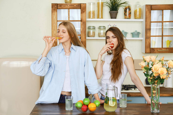 Two beautiful young women eat fruits in the kitchen - Foto, Imagen