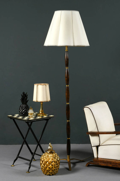 Décoration intérieure avec un mobilier vintage élégant comprenant un lampadaire en bois, chaise facile et lampe de table sur une table à damier avec des nuances de couleur crème et tapisserie contre un mur gris - Photo, image