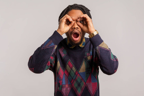 Extremely shocked surprised african guy with dreadlocks looking through fingers with opened mouth imagine binoculars, amazed with seen, spying. Indoor studio shot isolated on gray background - Photo, image