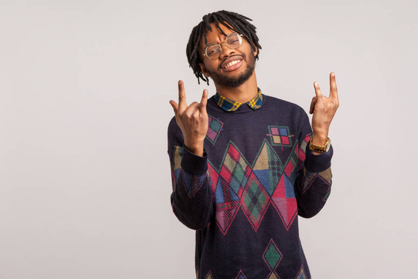 Naughty excited african man in eyeglasses with dreadlocks showing rock and roll hand gesture with toothy smile on face, cool and satisfied with life. Indoor studio shot isolated on gray background - Photo, Image