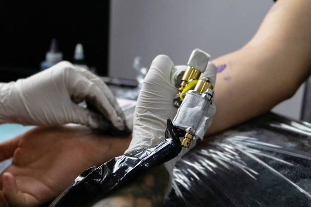 Ένας καλλιτέχνης τατουάζ γεμίζει ένα σχέδιο στο χέρι ενός ανθρώπου με μια γραφομηχανή. Η έννοια της υπέρβασης του πόνου, του θάρρους, της δημιουργικότητας και της σκέψης έξω από το κουτί - Φωτογραφία, εικόνα