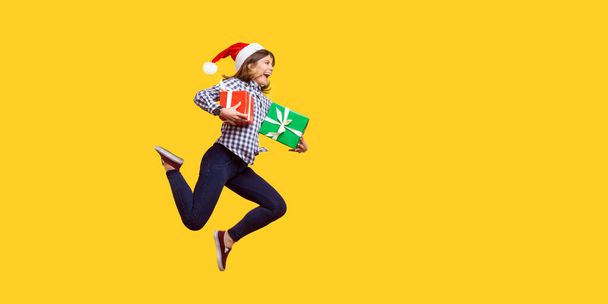 Side view portrait of extremely joyous brunette woman in santa hat jumping or running in air with wrapped xmas gift boxes, celebrating discounts. indoor studio shot isolated on yellow background - Fotó, kép