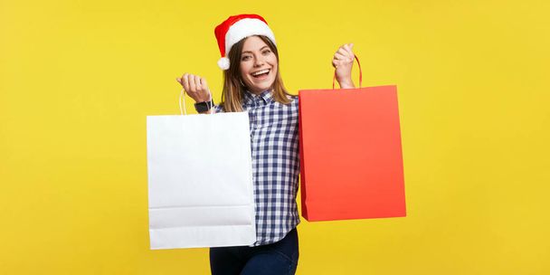 Porträt einer zufriedenen Käuferin, brünetten Frau mit Neujahrsmütze und charmantem Lächeln, die Einkaufstaschen zeigt und sich mit dem Kauf vor der Kamera rühmt. Innenstudio isoliert auf gelbem Hintergrund aufgenommen - Foto, Bild