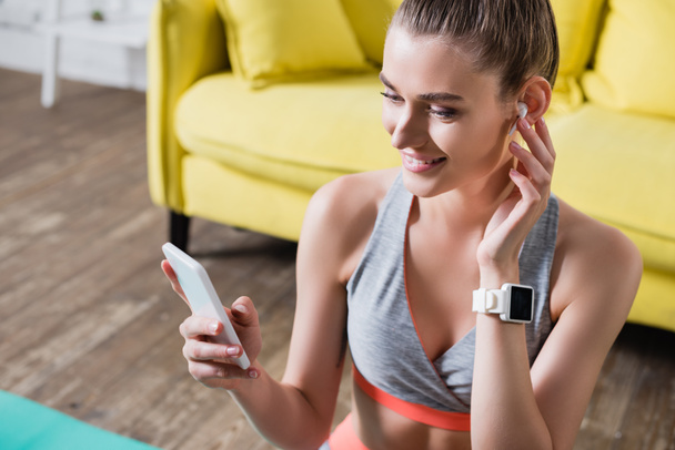 Sportswoman avec écouteur sans fil et smartwatch tenant le téléphone portable à la maison - Photo, image