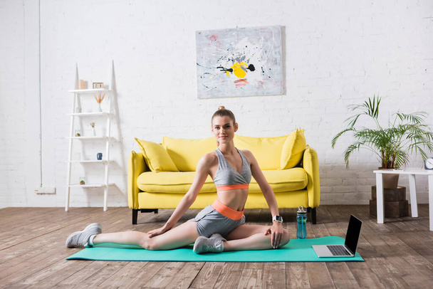 Sportswoman training near laptop and sports bottle on fitness mat at home - Фото, зображення