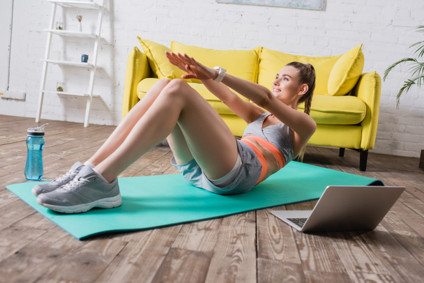 Selective focus of smiling sportswoman training on fitness mat near sports bottle and laptop  - Zdjęcie, obraz