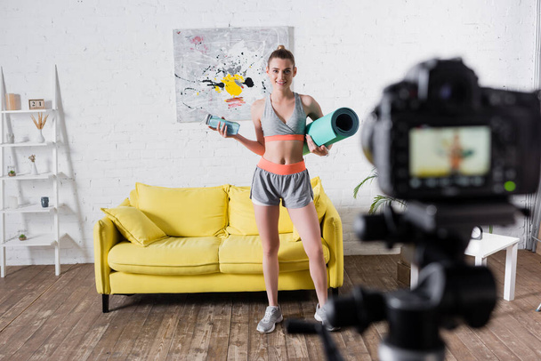 Selective focus of smiling sportswoman holding sports bottle and fitness mat near digital camera  - Photo, image