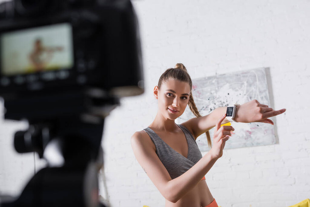 Selective focus of smiling sportswoman pointing at smart watch near digital camera at home  - Φωτογραφία, εικόνα
