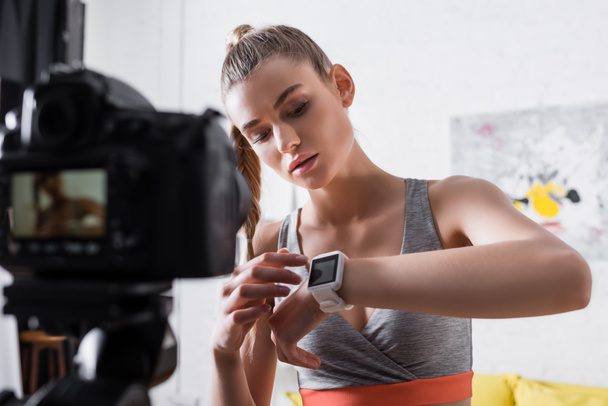 Enfoque selectivo de la mujer en ropa deportiva apuntando al rastreador de fitness cerca de la cámara digital en casa  - Foto, imagen
