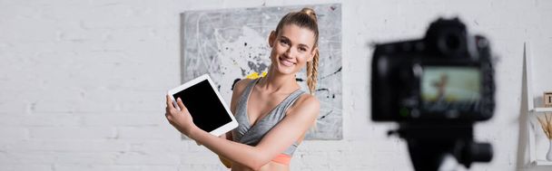 Panoramic shot of cheerful sportswoman holding digital tablet near digital camera in living room  - Valokuva, kuva