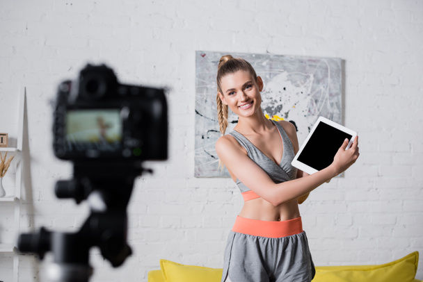 Enfoque selectivo de deportista positiva sosteniendo tableta digital con pantalla en blanco cerca de la cámara digital en casa  - Foto, imagen
