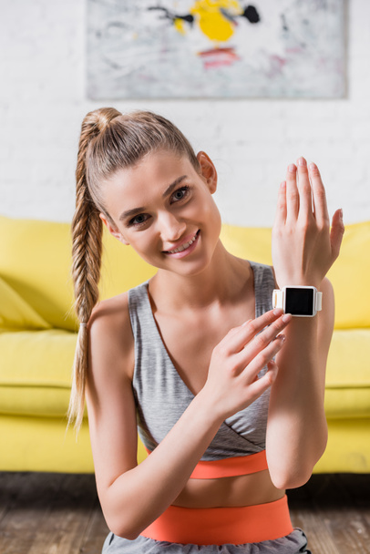 Smiling sportswoman touching fitness tracker and looking at camera at home - Foto, imagen