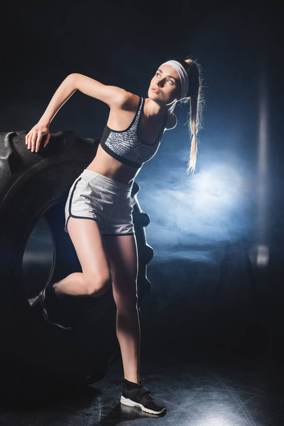Sportswoman in sportswear and headband exercising with tire during training in gym with smoke  - Фото, изображение