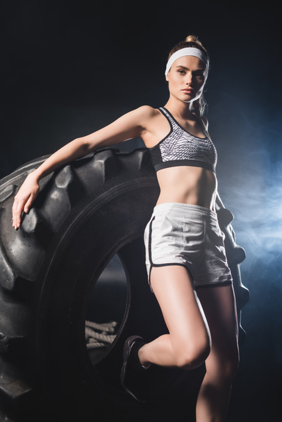 Foco seletivo da jovem mulher em sportswear e headband olhando para a câmera perto de pneu no ginásio com fumaça  - Foto, Imagem