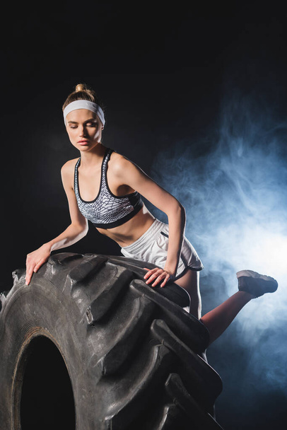 Selective focus of young sportswoman touching tire in gym with smoke  - Fotografie, Obrázek