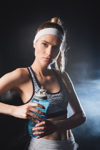 Young sportswoman holding sports bottle while looking at camera in gym with smoke  - Foto, Imagen