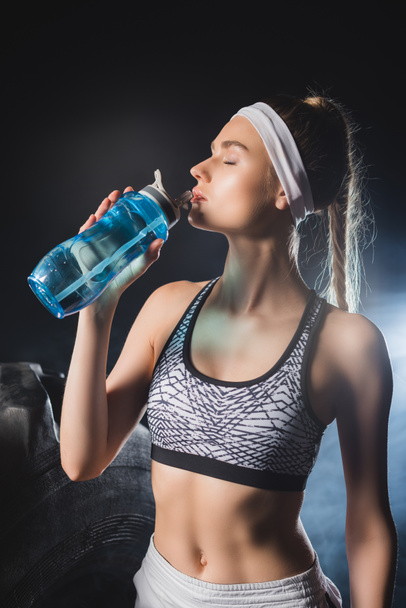 Sportswoman drinking water near tire in gym with smoke  - 写真・画像