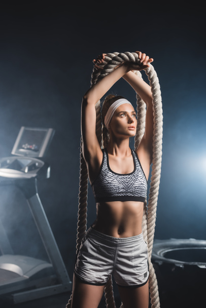 Sportswoman holding battle rope while standing in gym with smoke  - Photo, Image