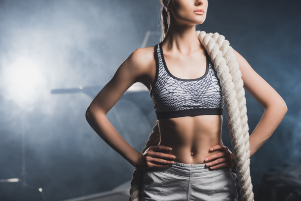 Cropped view of young sportswoman with battle rope and hands on hips standing in gym with smoke  - 写真・画像