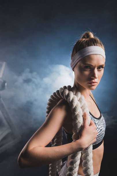 Young sportswoman holding battle rope while looking at camera in gym with smoke  - Fotografie, Obrázek
