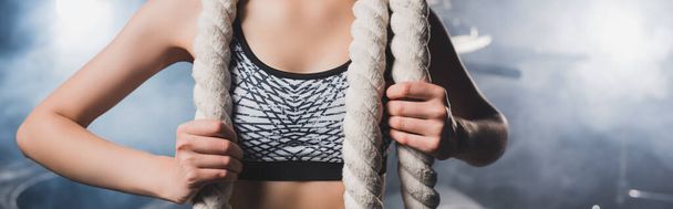 Panoramic shot of sportswoman holding battle rope near smoke in gym  - Photo, image