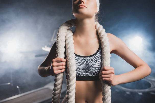 Cropped view of sportswoman in headband holding battle rope in gym with smoke  - Photo, Image