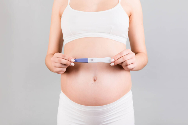 Close up of pregnant woman in white underwear holding positive pregnancy test at gray background. Motherhood concept. Copy space. - Фото, изображение