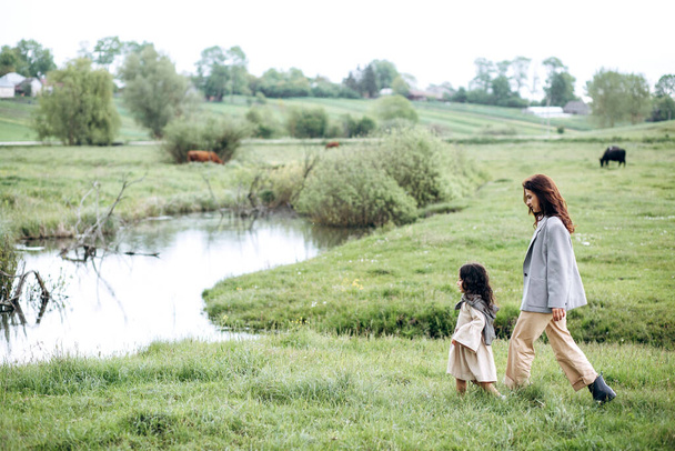 雨が降ると川のそばに緑の芝生が広がる野原で、スタイリッシュな母親と娘が楽しい時間を過ごしています。 - 写真・画像