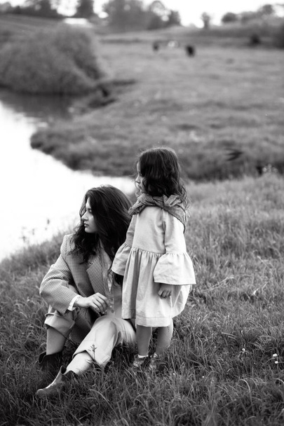 Elegante madre e hija se divierten al aire libre en un campo con hierba verde junto al río cuando llueve - Foto, imagen