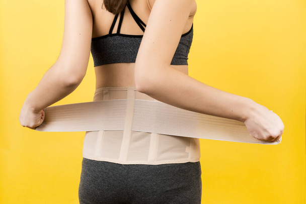 Back view of pregnant woman dressing orthopedic corset to make the backache go away at yellow background with copy space. Close up of orthopedic abdominal support belt concept. - Photo, Image