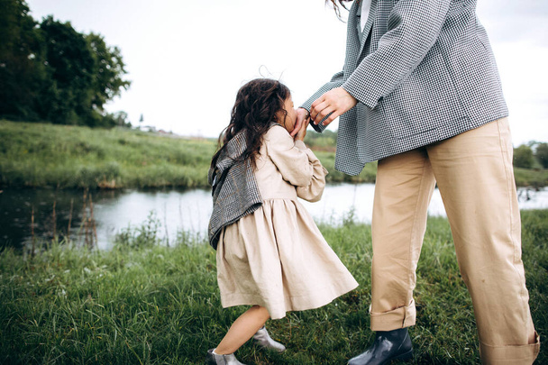 Stylish mother and daughter have fun outdoors in a field with green grass by the river when it rains - Photo, Image