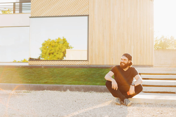 Genç sakallı bir erkek gülümsüyor ve evinin yakınına bakıyor. - Fotoğraf, Görsel