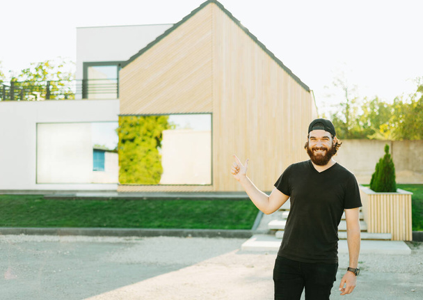 Bärtiger junger Mann blickt in die Kamera und zeigt auf sein neues Haus im Hintergrund - Foto, Bild